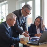 Foto de reunião entre dois homens (um de pé e um sentado) e uma mulhersentada frente a um computador