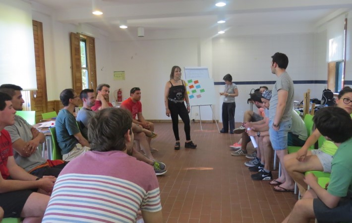 Grupo de pessoas sentadas numa sala, a rodear a formadora de pé. Momento de debriefing.